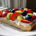 A close-up of a slice of fruit pizza sugar cookie topped with cream cheese frosting, strawberries, blueberries, and kiwi.
