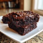 Two fudgy, dark chocolate sweet potato brownies stacked on a white plate, topped with chocolate chips.
