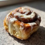 A close-up, appetizing photo of a single, freshly baked cinnamon roll topped with thick, dripping white icing.