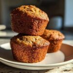 Three golden-brown banana protein muffins stacked on a white plate, highlighted by dramatic natural light.