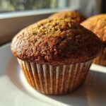 Close-up of a freshly baked, dark golden honey bran muffin sitting on a white plate with two others blurred in the background.