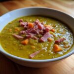 Close-up of a thick, yellow-green pea soup topped with diced ham and carrots in a white bowl.