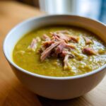 Close-up of a white bowl filled with thick, green pea soup, garnished generously with shredded ham.