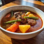 Close-up of a steaming bowl of rich vegetable beef soup featuring large chunks of beef, potatoes, carrots, and green beans.
