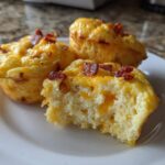 Three fluffy, high-protein egg bites topped with melted cheddar and bacon bits, one is bitten into, showing the texture of the egg bites.