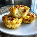 Three fluffy, golden-brown high-protein egg bites, one broken open, showing bacon pieces, served on a white plate.
