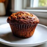 A single, dark golden brown honey bran muffin sitting on a white plate near a window.