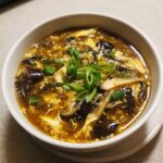 A close-up of a steaming bowl of hot and sour soup recipe, featuring dark broth, shredded egg, mushrooms, and green onion garnish.