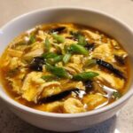 A close-up of a white bowl filled with steaming hot and sour soup recipe, featuring egg ribbons and green onions.