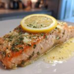 Close-up of a perfectly cooked piece of baked salmon recipe topped with garlic, parsley, and a lemon slice.