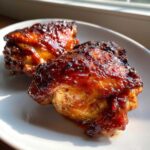 Two pieces of incredibly juicy bbq chicken with a dark, caramelized glaze resting on a white plate.