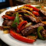 Close-up of perfectly cooked steak fajitas featuring strips of seasoned beef mixed with vibrant red and green bell peppers and onions.