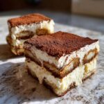 A close-up of a rich, layered slice of tiramisu recipe, dusted heavily with cocoa powder.