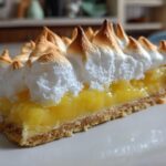 A close-up slice of lemon meringue pie showing bright yellow curd, crust, and perfectly peaked, toasted meringue.