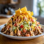 A tall, appetizing serving of taco salad piled high with seasoned ground beef, lettuce, tomatoes, olives, cheese, and tortilla chips.