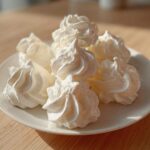A close-up of several perfectly piped, white meringue cookies stacked on a small white plate.