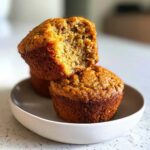 Two golden brown banana protein muffins stacked, one broken open to show the moist interior texture.