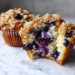 Close-up of a moist blueberry muffin cut open, showing rich purple berries and a crunchy streusel topping.