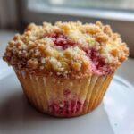 Close-up of a moist strawberry muffin with a golden crumb topping, showing bright pink fruit inside.