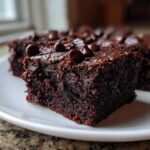 Close-up of a rich, dark chocolate sweet potato brownies slice topped with chocolate chips on a white plate.