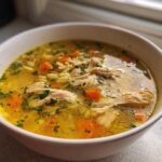 A close-up of a white bowl filled with golden penicillin soup, featuring shredded chicken, carrots, and herbs.