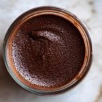 Overhead view of rich homemade chocolate pudding with a slightly textured surface in a small glass jar.