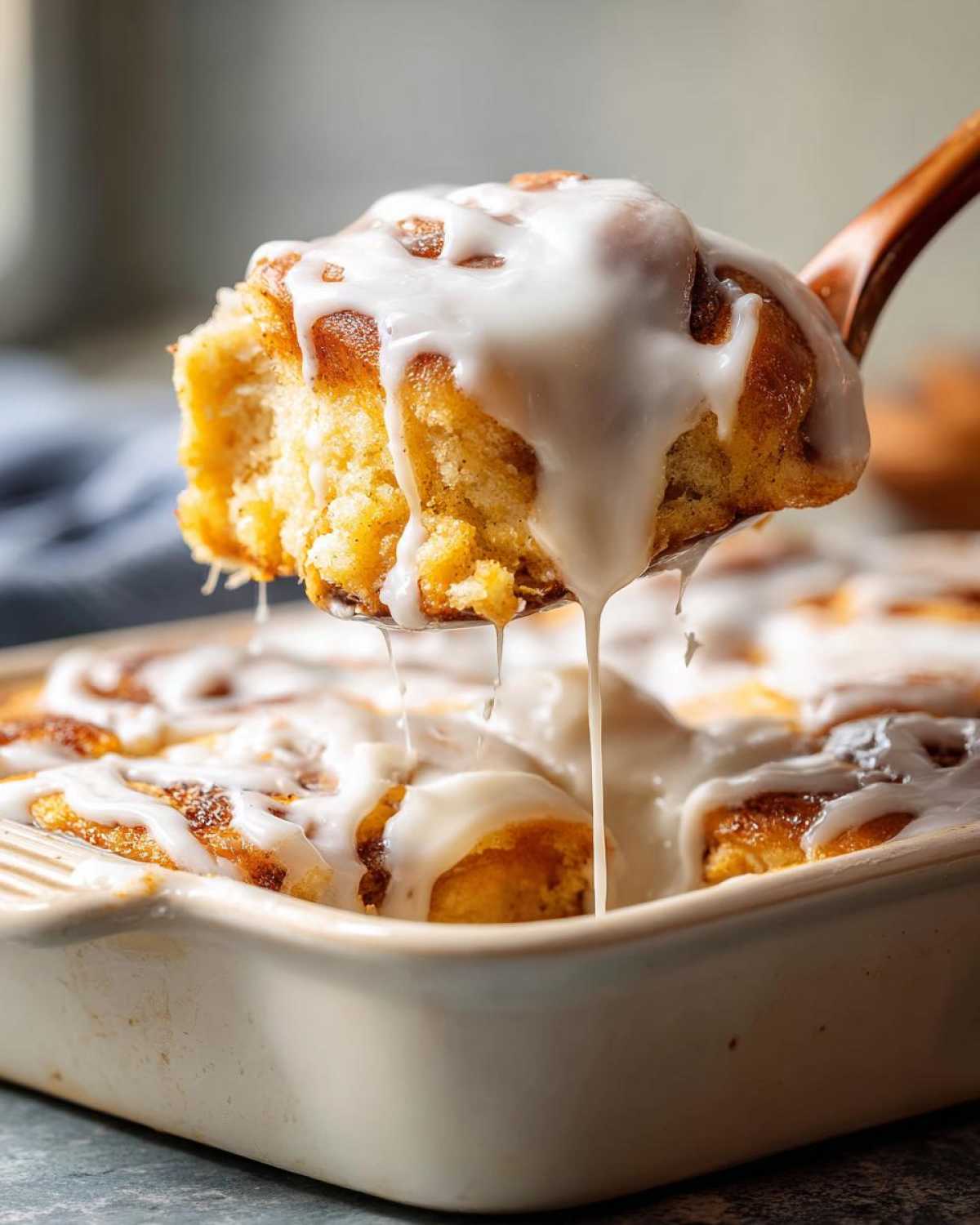 A spoonful of gooey Cinnamon roll casserole topped with thick white icing dripping back into the baking dish.
