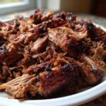 Close-up of tender, shredded Slow Cooker Pulled Pork piled high on a white plate, showing caramelized edges.