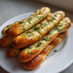 A stack of freshly baked, soft garlic butter breadsticks topped generously with chopped parsley on a white plate.