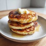 A stack of three golden-brown sourdough pancakes drizzled with syrup and topped with a dollop of melting butter.