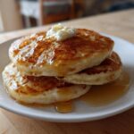 A close-up of three fluffy sourdough pancakes stacked on a white plate, topped with melting butter and drizzled with syrup.