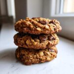 A stack of three perfectly baked, chewy oatmeal cookie with melted chocolate chips on a white marble surface.