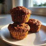 Three freshly baked, dark brown bran muffins stacked and resting on a light plate, highlighted by warm sunlight.