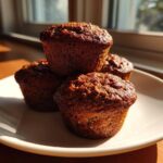 Four dark, moist bran muffins stacked on a white plate, illuminated by strong sunlight from a window.