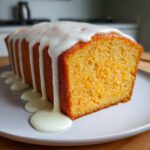 A close-up of a moist, orange-flecked sweet potato pound cake loaf topped with thick white glaze dripping down the sides.