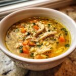 Close-up of a white bowl filled with steaming penicillin soup, featuring shredded chicken, diced carrots, orzo pasta, and fresh parsley.