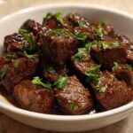 Close-up of tender, saucy crockpot steak bites garnished with fresh parsley in a white bowl.