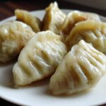 Close-up of several perfectly pleated Traditional Chinese Dumplings, lightly browned on the bottom, served on a white plate.