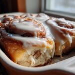 Close-up of freshly baked, soft cinnamon roll covered in thick white icing, sitting in a baking dish.