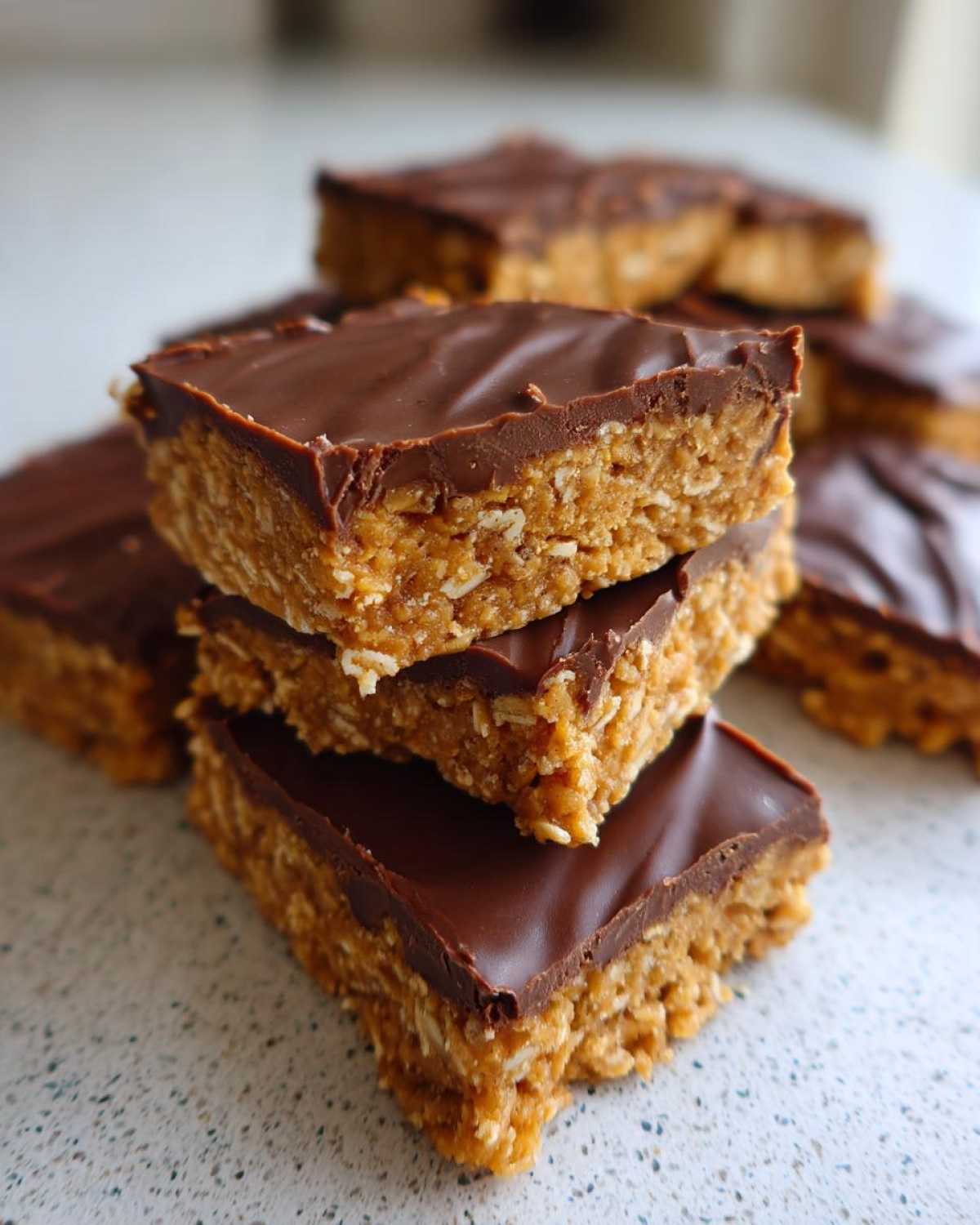 A stack of homemade, no-bake protein bars featuring an oat base and a thick chocolate topping.