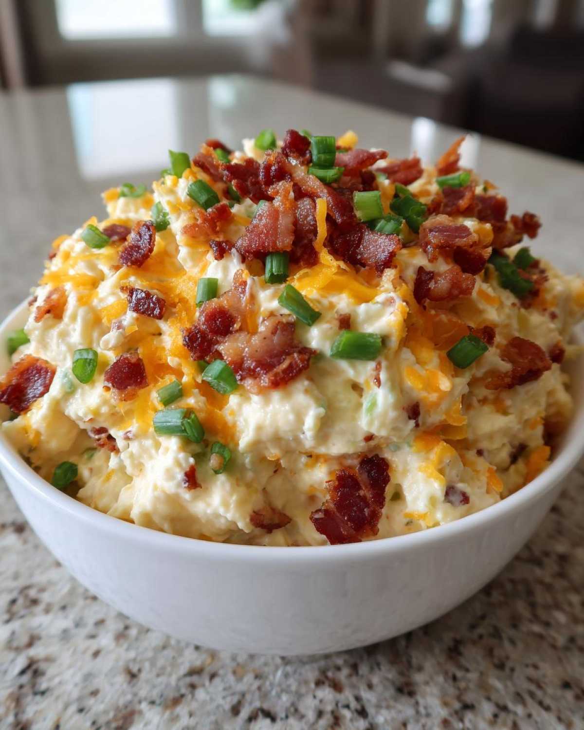 A white bowl filled with creamy Million Dollar Dip, generously topped with shredded cheddar cheese, crispy bacon bits, and fresh green onions.