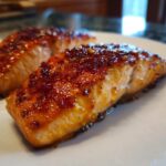 Close-up of two perfectly cooked salmon fillets with a shiny, caramelized glaze on a white plate.