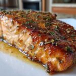 A close-up of a perfectly cooked, glazed salmon fillet topped with fresh herbs resting in its juices.
