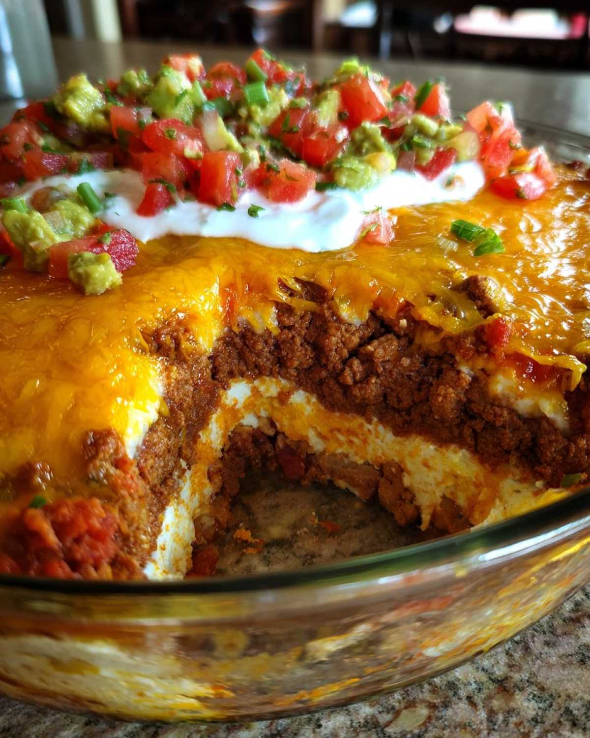 Close-up view showing the layers of a 7 layer dip in a glass dish, topped with melted cheese, sour cream, and fresh salsa/guacamole.