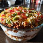 A vibrant, tall 7 layer dip served in a clear glass bowl, topped with shredded cheese, tomatoes, and guacamole.