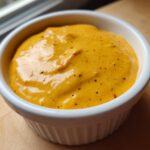 Close-up of a thick, vibrant orange beer cheese dip speckled with spices, served in a white ramekin.