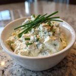 Close-up of a chunky, creamy blue cheese dip in a white bowl, garnished with fresh green chives.