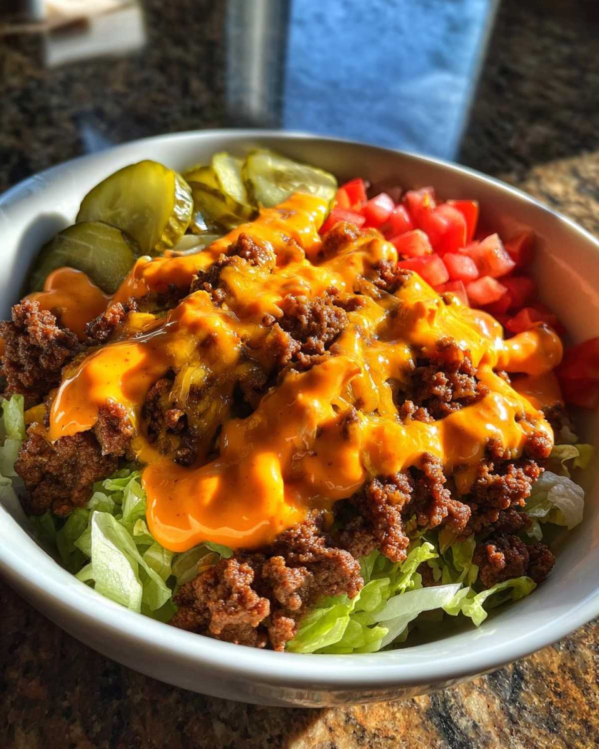 A close-up of an amazing cheeseburger bowl featuring ground beef, melted cheese sauce, lettuce, pickles, and diced tomatoes.