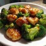 Close-up of glossy, sauced chicken pieces mixed with bright green broccoli florets in a white bowl.