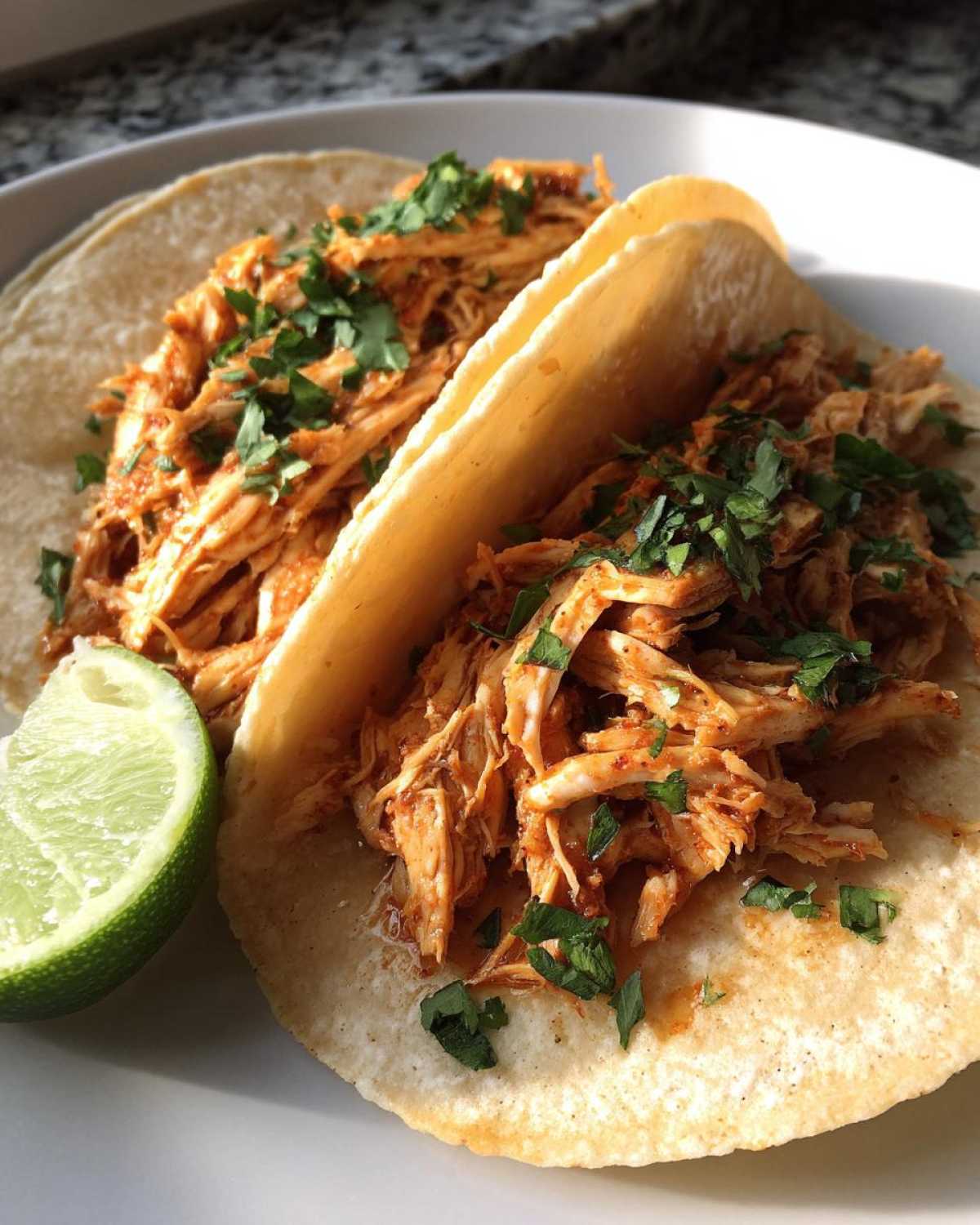 Two soft corn tortillas filled with shredded, seasoned chicken tacos, topped with fresh cilantro and a lime wedge.